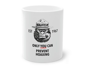 May include: White ceramic mug featuring a Bigfoot-like character wearing a ranger hat. The mug has the text "SQUATCHY EST 1967" and "ONLY YOU CAN PREVENT HOAXING" in black lettering.