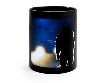 May include: Black ceramic mug featuring a silhouette of a large, hairy creature in front of a car with bright headlights. The background is a dark blue, creating a mysterious and eerie scene. The mug is a standard 11 oz size.