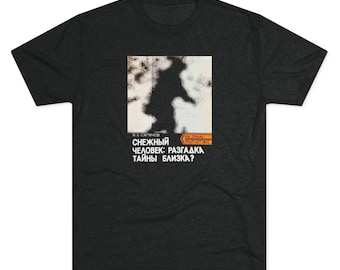 May include: A charcoal gray short-sleeve t-shirt featuring a central graphic print. The design includes a pixelated black silhouette of a figure against a white background, with Russian text reading "СНЕЖНЫЙ ЧЕЛОВЕК: РАЗГАДКА ТАЙНЫ БЛИЗКА?" and an orange rectangular accent.