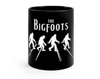 May include: Black ceramic mug featuring the text "The Bigfoots" in white, above a silhouette of four Bigfoot figures walking on stilts. The mug has a glossy finish and a simple, cylindrical shape, suitable for hot beverages.