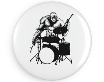 May include: A white button featuring a black and white illustration of a Bigfoot playing a drum set. The Bigfoot is depicted with a muscular build and is holding drumsticks. The drum set includes a bass drum, snare drum, and cymbals.