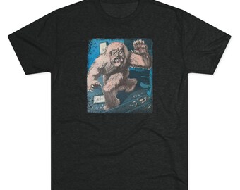 May include: Dark gray t-shirt featuring a vintage-style illustration of a Bigfoot creature. The graphic shows a hairy, light brown Bigfoot with a menacing expression, set against a blue and gray background. The design is printed on a soft, comfortable tee.