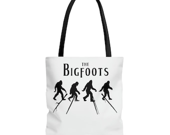 May include: White tote bag with black handles featuring the text "THE BIGFOOTS" above a silhouette of four Bigfoot creatures walking on stilts. The design is in black and white.