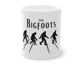 May include: White ceramic mug featuring the text "THE BIGFOOTS" above a silhouette of four Bigfoot figures walking on stilts. The black figures are in a row, with the stilts angled. A fun, novelty item.