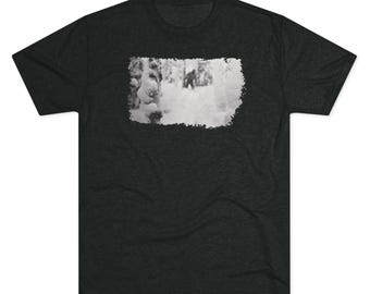 May include: Dark gray t-shirt featuring a black and white image of a snowy forest scene. The image is framed with a white border. The shirt has a crew neck and short sleeves.