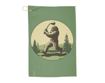 May include: A green golf towel with a circular design featuring a brown Bigfoot swinging a golf club. The Bigfoot is in a forest setting with green trees.