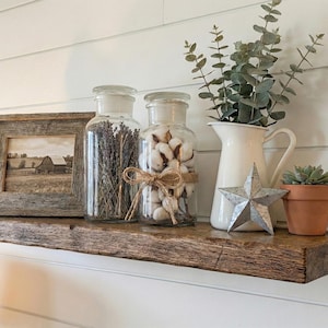 May include: A rustic wooden shelf displays a framed sepia-toned picture of a barn, glass jars filled with lavender and cotton, a white pitcher with greenery, a metal star, and a small potted succulent. The shelf is made of reclaimed wood.