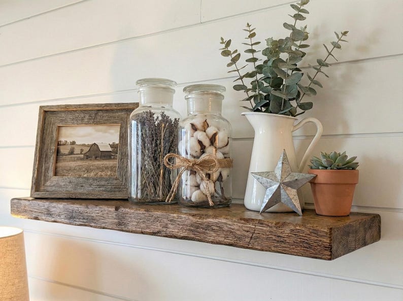 K&ouml;nnte beinhalten: Ein rustikales Holzregal zeigt ein gerahmtes Sepia-Foto einer Scheune, Gl&auml;ser mit Lavendel und Baumwolle, einen wei&szlig;en Krug mit Gr&uuml;nzeug, einen Metallstern und eine kleine Topf-Sukkulente. Das Regal ist aus Altholz.