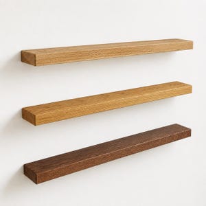May include: Three floating wooden shelves in varying shades of brown. The top shelf is light oak, the middle shelf is medium oak, and the bottom shelf is a dark walnut color. Each shelf is rectangular and mounted on a white wall.