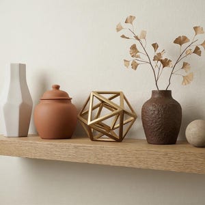 May include: A wooden shelf displays decorative items. A white geometric vase, a terracotta jar with a lid, a gold geometric sculpture, a textured brown vase with dried branches, and a stone sphere are arranged on the shelf.