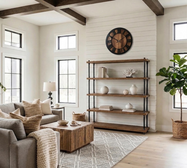 May include: A large round wall clock with a dark black frame and a brown wood-grain face featuring black Roman numerals and thin black hands, mounted on a white shiplap wall above a four-tier open shelving unit made of dark metal pipes and wood.