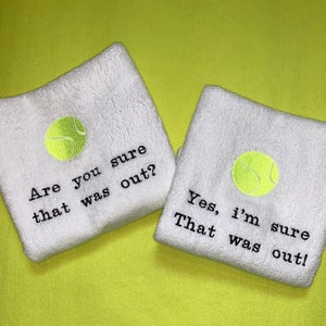 Könnte beinhalten: Zwei weiße Handtücher mit gestickten Tennisbällen und Text. Das erste Handtuch sagt "Sind Sie sicher, dass das draußen war?" Das zweite Handtuch sagt "Ja, ich bin sicher. Das war draußen!"