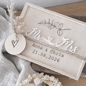Puede incluir: Un letrero de boda de madera con las palabras "Mr & Mrs" en escritura blanca, los nombres "Anna & Chris" y la fecha "21.06.2026". El letrero está decorado con un diseño floral y un pequeño círculo de madera con un corazón.