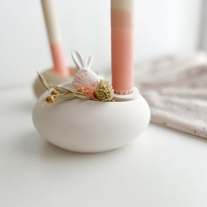 Może przedstawiać: Biały ceramiczny świecznik z motywem uszu królika, trzymający różowo-białą świecę ombre. Uchwyt jest ozdobiony suszonymi kwiatami i zielenią. W tle druga świeca i uchwyt.
