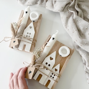 Könnte beinhalten: Zwei weiße Keramikhäuser mit schwarzen Herzdetails, jedes mit einer weißen Kerze und einem kleinen Holzsockel. Die Häuser sind mit Schnur gebunden und mit getrockneten Blumen verziert.