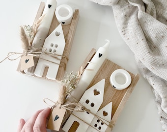 May include: Two white ceramic houses with black heart details, each with a white candle and a small wooden base. The houses are tied with twine and decorated with dried flowers.