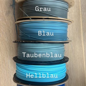 May include: Spools of 3D printing filament in various colors, including black, gray, and shades of blue. Each spool is labeled with its color name. The image shows a variety of filament options for 3D printing projects.