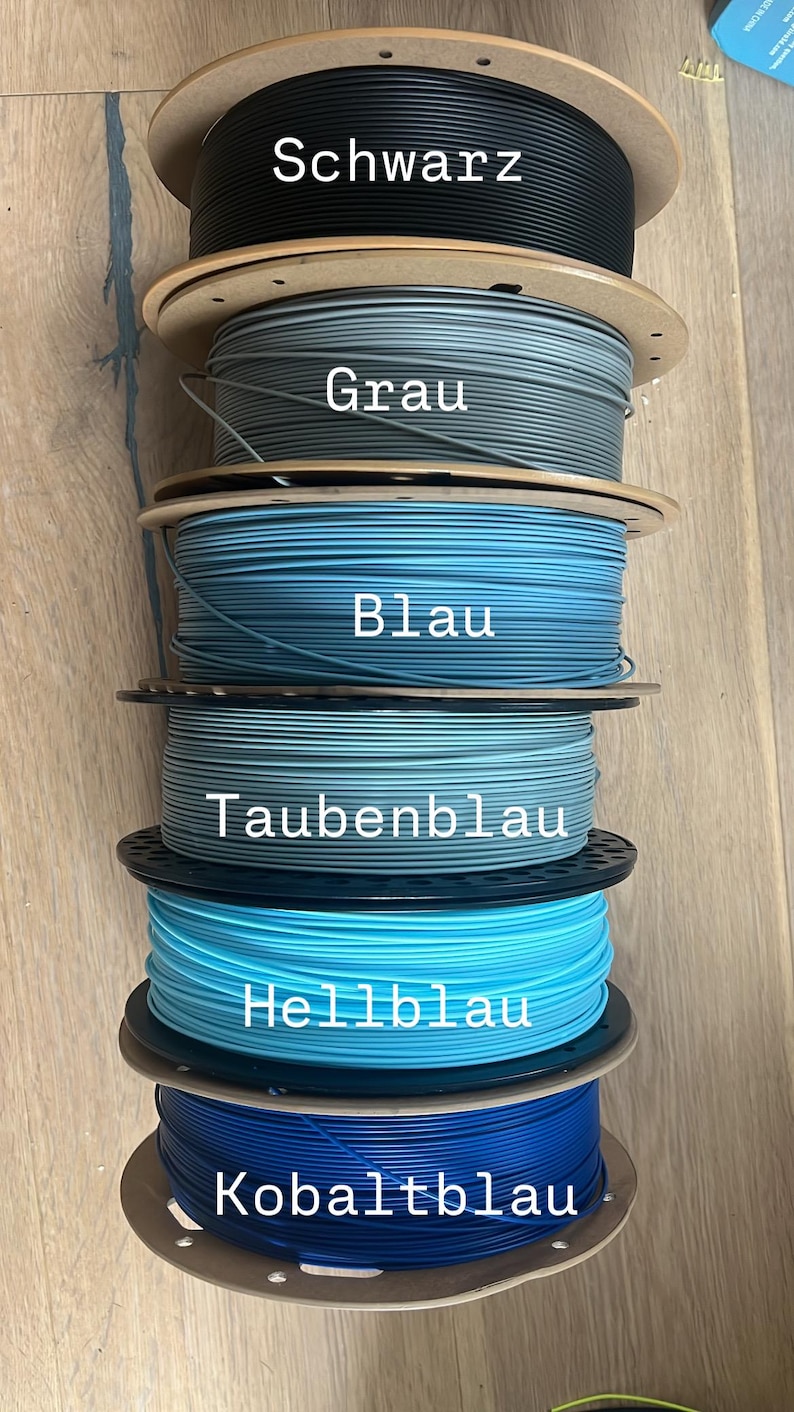 May include: Spools of 3D printing filament in various colors, including black, gray, and shades of blue. Each spool is labeled with its color name. The image shows a variety of filament options for 3D printing projects.