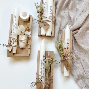 May include: Four wooden boxes with candles and dried flowers. The candles are white and have text on them. The boxes are tied with twine and have small tags with hearts on them. The text on the candles reads "Danke, dass du mir beim Wachsen geholfen hast", "Janke HDMI", "We", and "STATT Blumen".
