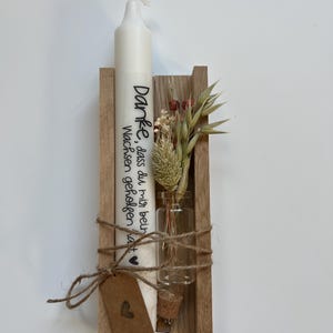 May include: A white candle with the text "Danke, dass du mir beim Wachsen geholfen hast" in black lettering is displayed in a wooden holder with a small glass vial containing dried flowers.