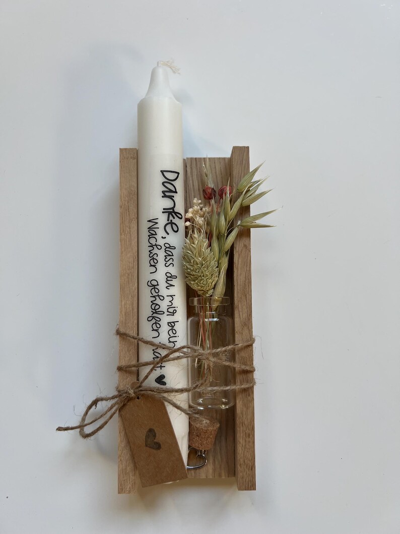 May include: A white candle with the text "Danke, dass du mir beim Wachsen geholfen hast" in black lettering is displayed in a wooden holder with a small glass vial containing dried flowers.