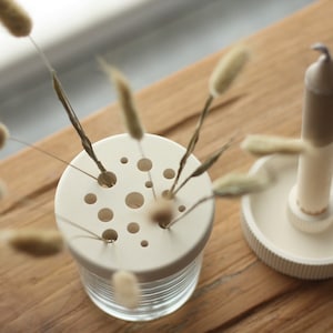 May include: A small, clear glass vase with a white, perforated lid, holding dried flowers. A white candle sits in a matching holder. The items are on a wooden surface, creating a minimalist, natural aesthetic.