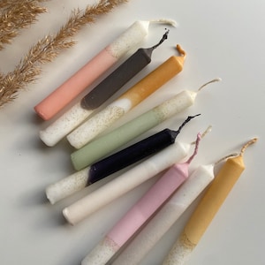 Könnte beinhalten: Eine Sammlung von acht Stumpenkerzen mit einem zweifarbigen Design. Die Kerzen sind weiß mit einer zweiten Farbe auf der unteren Hälfte, darunter rosa, grün, gelb, schwarz und grau.
