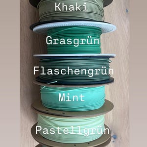 May include: A stack of spools of 3D printing filament in various shades of green, including neon, khaki, mint, and sage. Each spool is labeled with its color name in white text. The spools are stacked on top of each other.