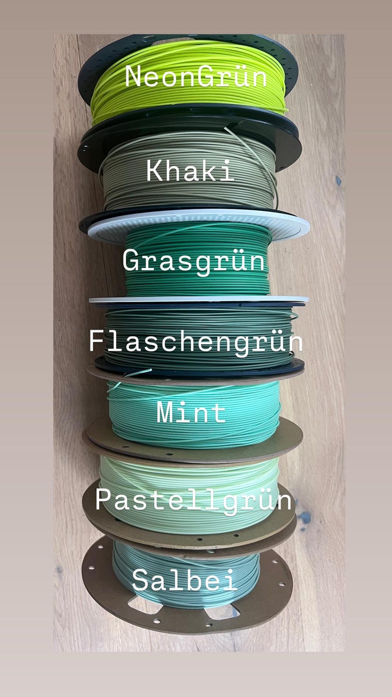 May include: A stack of spools of 3D printing filament in various shades of green, including neon, khaki, mint, and sage. Each spool is labeled with its color name in white text. The spools are stacked on top of each other.
