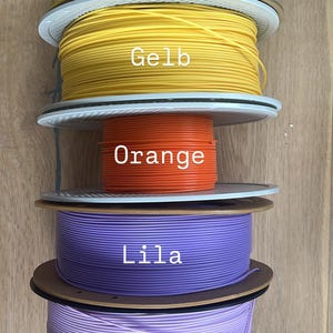 May include: A stack of colorful 3D printing filament spools. The colors include neon yellow, yellow, orange, purple, and lilac. Each spool has a label with the color name. The spools are on a wooden surface.