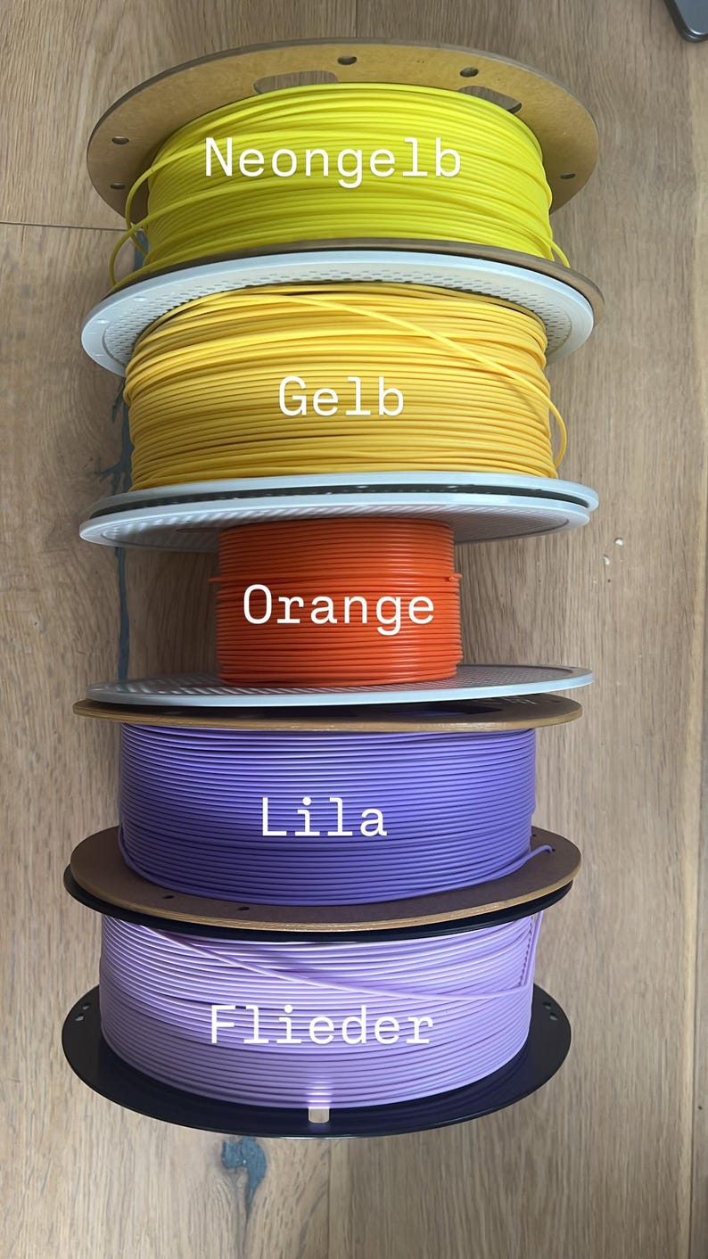 May include: A stack of colorful 3D printing filament spools. The colors include neon yellow, yellow, orange, purple, and lilac. Each spool has a label with the color name. The spools are on a wooden surface.