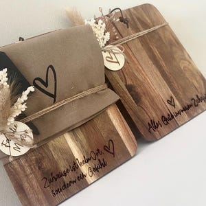 Puede incluir: Dos tablas de cortar de madera con elementos decorativos. Una tabla tiene un corazón y texto, la otra texto. Se incluye un paquete envuelto con un diseño de corazón y flores secas. Las tablas son marrones con vetas de madera natural.