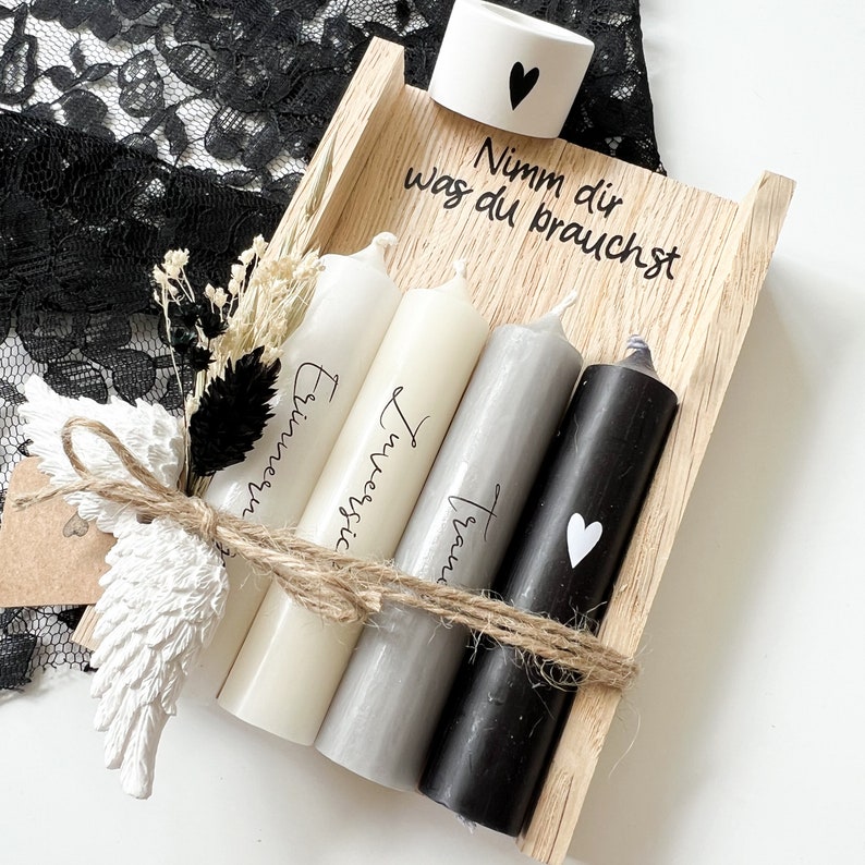 May include: A wooden box with four candles inside. The candles are white, gray, and black. The box has the text "Nimm dir was du brauchst" written on it. The candles are tied together with twine and a small white angel wing ornament.