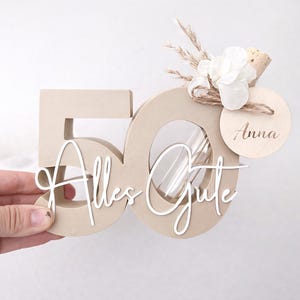 May include: A beige wooden '50' shaped decoration with the words 'Alles Gute' in white script. It is adorned with a small bouquet of white flowers, dried grass, and a wooden tag that says 'Anna'.