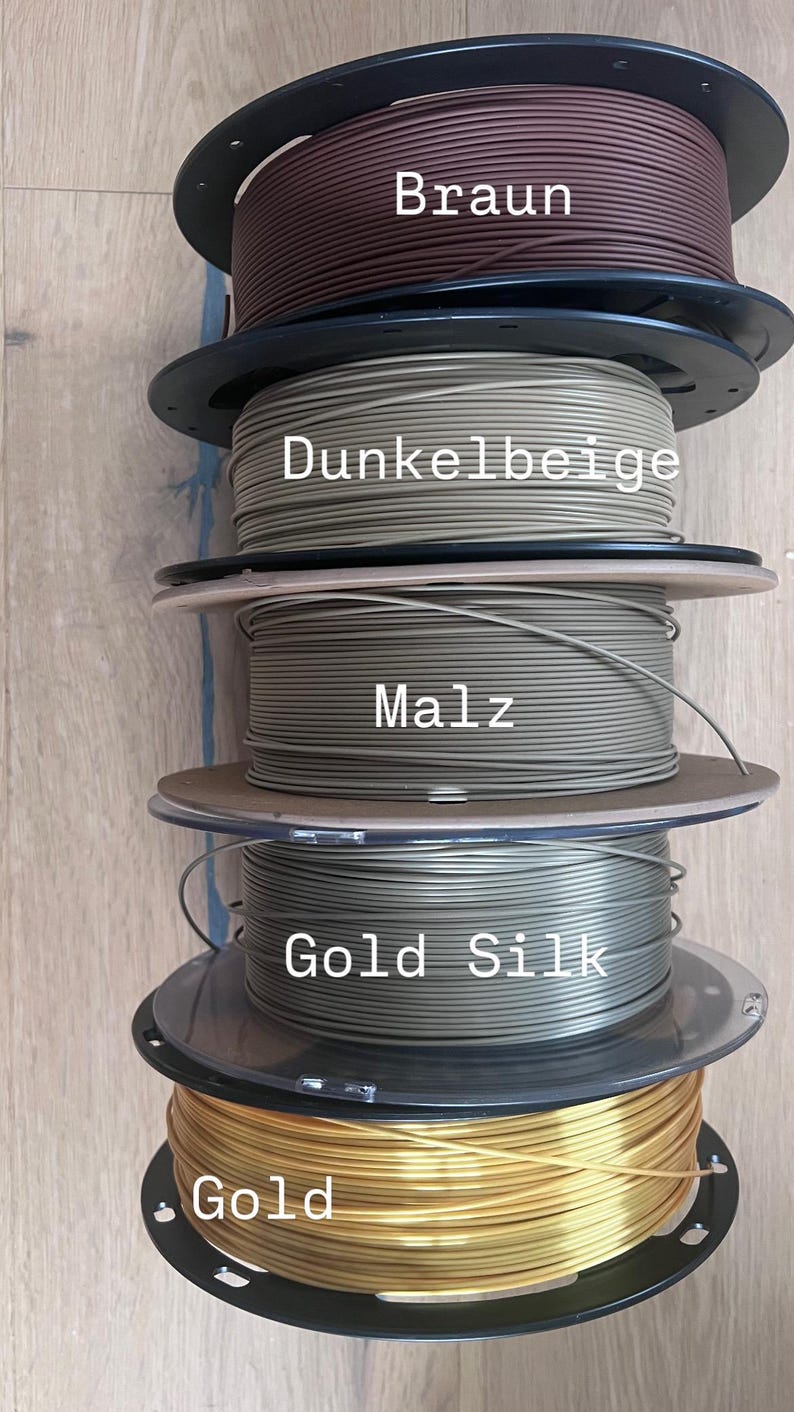 May include: A stack of five spools of 3D printing filament in various colors. The colors include brown, dark beige, malz, gold silk, and gold. Each spool is labeled with its color name.