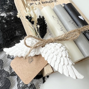 May include: A wooden tray with four candles, a small white angel wing, and a tag with a heart. The candles are white, gray, and black. The tray has the text "Nimm dir was du brauchst" written on it.