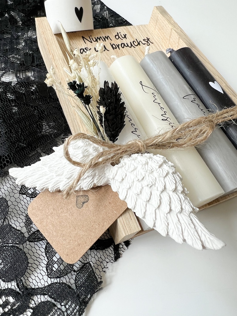 May include: A wooden tray with four candles, a small white angel wing, and a tag with a heart. The candles are white, gray, and black. The tray has the text "Nimm dir was du brauchst" written on it.