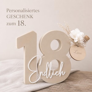May include: A beige wooden number 18 with the word "Endlich" in white script. A small glass bottle with a cork, flowers, and a wooden tag that says "Lena" is attached to the number 8. The text "Personalisiertes GESCHENK zum 18." is at the top.