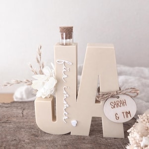 Könnte beinhalten: Beige Holzskulptur in Form der Buchstaben "JA" mit einer kleinen Glasflasche und einem Korken. Der Text "für immer" ist vertikal auf dem "J" geschrieben. Ein Holzschild mit "SARAH & TIM" ist angebracht.