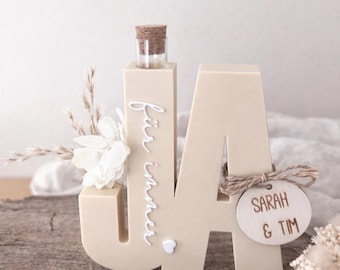 Könnte beinhalten: Beige Holzskulptur in Form der Buchstaben "JA" mit einer kleinen Glasflasche und einem Korken. Der Text "für immer" ist vertikal auf dem "J" geschrieben. Ein Holzschild mit "SARAH & TIM" ist angebracht.