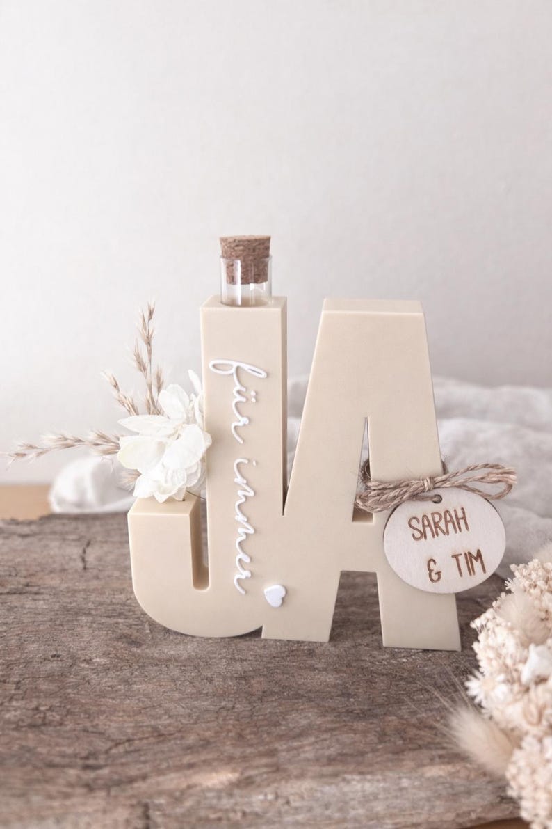 Puede incluir: Escultura de madera beige con forma de letras "JA" con una peque&ntilde;a botella de vidrio y un tap&oacute;n de corcho. El texto "f&uuml;r immer" est&aacute; escrito verticalmente en la "J". Una etiqueta de madera con "SARAH & TIM" est&aacute; adjunta.