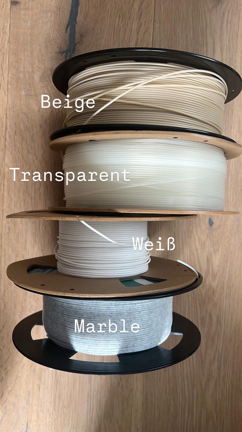May include: Four spools of 3D printer filament in various colors: beige, transparent, white, and marble. Each spool is wound on a cardboard and black plastic reel. The colors are labeled on the image.