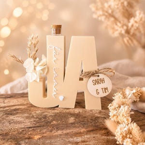 Puede incluir: Decoraci&oacute;n de boda de madera beige 'UA' con una peque&ntilde;a botella de vidrio y corcho. La letra 'U' tiene flores secas y el texto 'f&uuml;r immer' en cursiva. Una etiqueta de madera dice 'SARAH & TIM'.