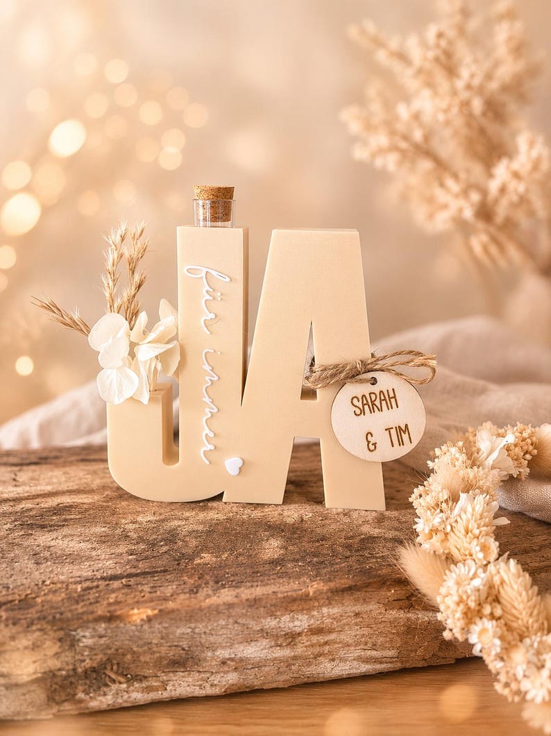 Puede incluir: Decoraci&oacute;n de boda de madera beige 'UA' con una peque&ntilde;a botella de vidrio y corcho. La letra 'U' tiene flores secas y el texto 'f&uuml;r immer' en cursiva. Una etiqueta de madera dice 'SARAH & TIM'.