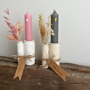 May include: Two white ceramic candle holders with heart details, each holding a candle and dried flowers. The candles are pink and grey with a small heart detail on each. The holders have brown paper tags with the text "Von Herzen" on them.
