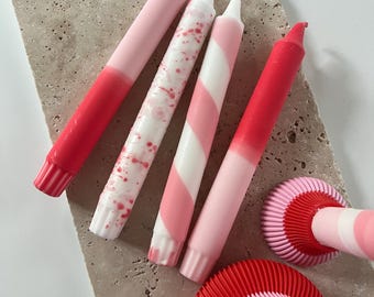 May include: A collection of decorative candles in shades of pink and red. The candles feature various designs, including solid colors, splattered patterns, and striped patterns. A matching candle holder is also visible.