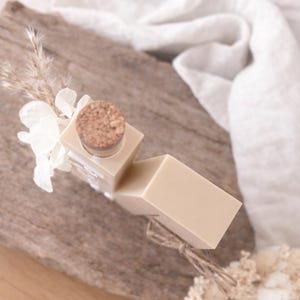 Puede incluir: Una pastilla de jab&oacute;n beige claro y una botella peque&ntilde;a con tap&oacute;n de corcho est&aacute;n dispuestas sobre una superficie de madera. La botella est&aacute; decorada con flores blancas y plantas secas. Un pa&ntilde;o blanco est&aacute; en el fondo.