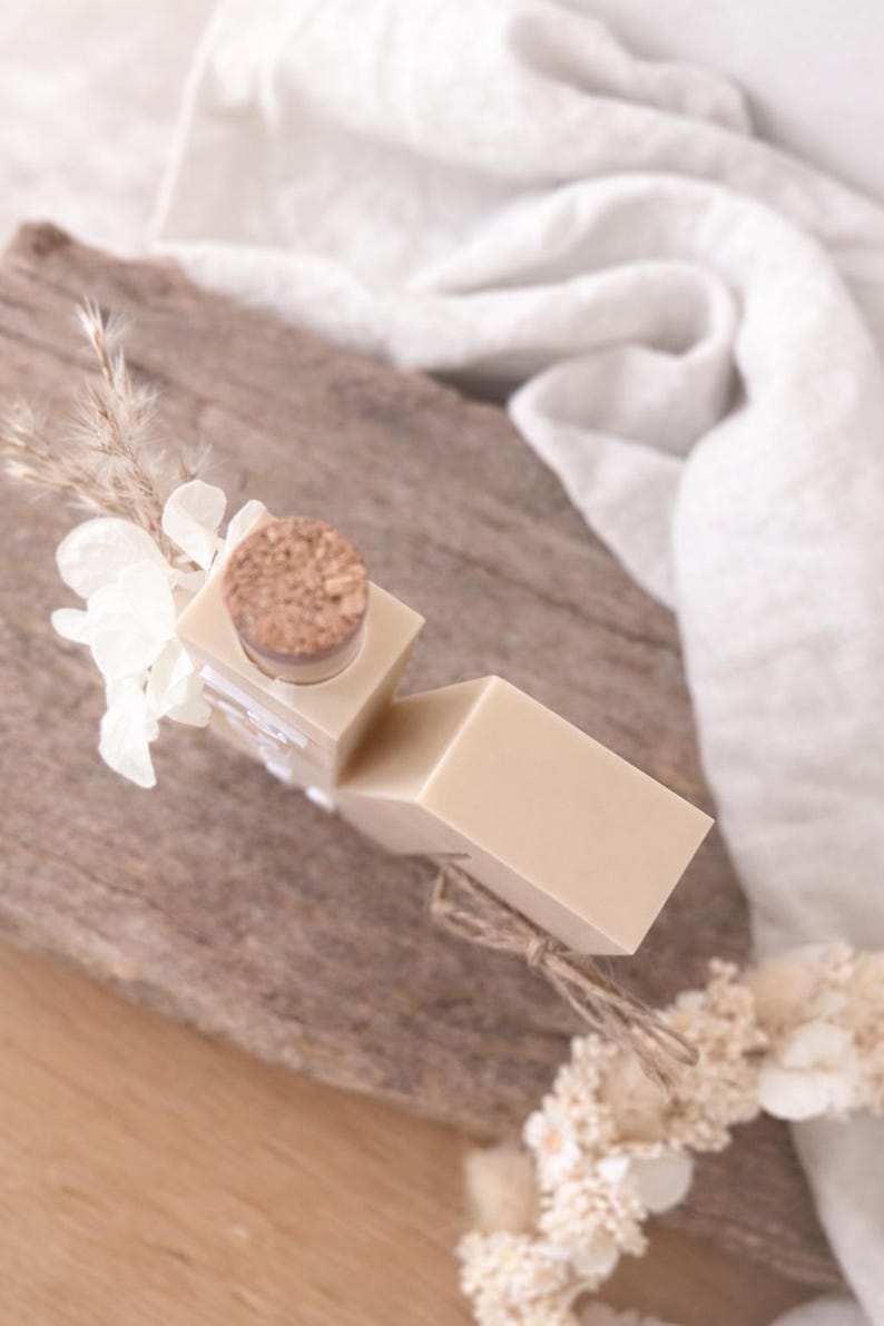 Puede incluir: Una pastilla de jab&oacute;n beige claro y una botella peque&ntilde;a con tap&oacute;n de corcho est&aacute;n dispuestas sobre una superficie de madera. La botella est&aacute; decorada con flores blancas y plantas secas. Un pa&ntilde;o blanco est&aacute; en el fondo.