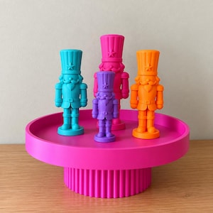 May include: Four nutcracker figurines in bright neon colors stand on a circular, ribbed, hot pink pedestal. The figurines are teal, magenta, purple, and orange, each featuring a tall hat with a star emblem and a mustache.