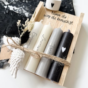May include: A wooden box with four candles inside. The candles are white, gray, and black. The box is tied with twine and has a small white heart on the side. The text on the box reads "Nimm dir was du brauchst".